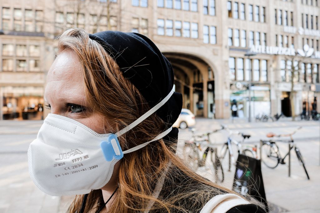 Seitenansicht einer jungen Frau mit langen, rotblonden Haaren, schwarzer Mütze und FFP3 Maske. Im hintergrund verschwommen helle Häuser und Fahrräder