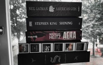 Eine Hand hält einen Stapel Bücher hoch (von unten nach oben): House of Leaves, The Ship of Theseus, Aera, Shining, American Gods