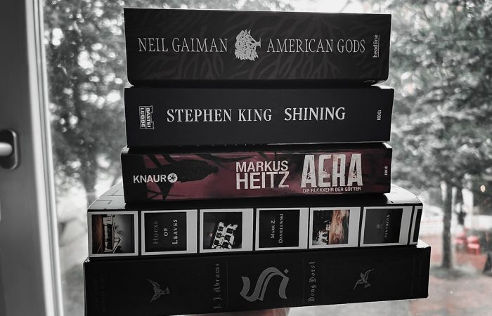Eine Hand hält einen Stapel Bücher hoch (von unten nach oben): House of Leaves, The Ship of Theseus, Aera, Shining, American Gods