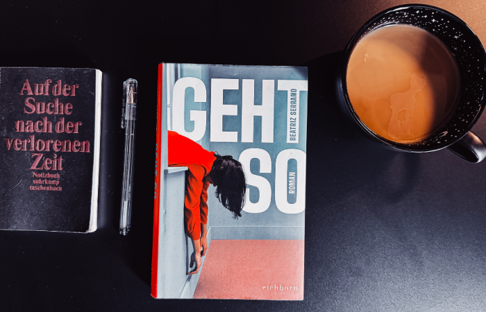 Links ein schwarzes Notizbuch mit Stift, in der Mitte das Buch "Geht so" von Beatriz Serrano, rechts eine schwarze Tasse mit Milchkaffee
